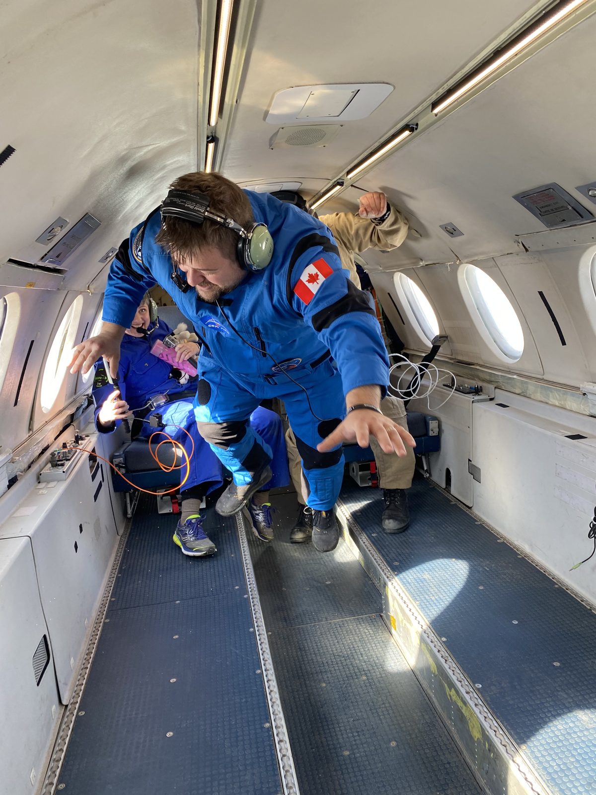 Jason Michaud in a blue flight suit, floating in microgravity on a parabolic flight, Canada flag on his shoulder